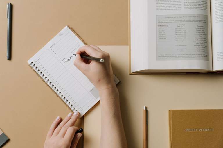 Hands writing in a weekly planner beside an open book.